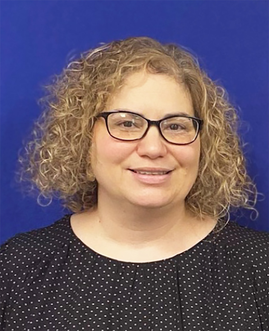 A portrait headshot photograph of Trisha Rosokoff (Buffalo Teachers Foundation) smiling with black glasses in a black and white polkadot pattern style shirt in front of a blue background