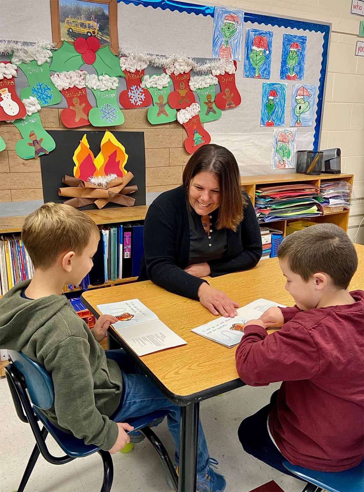 A portrait photograph of Laura Rosner in a black jacket (who is a member of the East Hampton Teachers Association and a co-teacher in an integrated first grade classroom) smiling and sitting with her two students (one wearing a green jacket and the other a red sweatshirt) discussing classwork