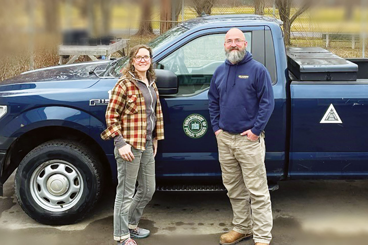 City Executive Association president Jeanne Grace (wearing a tan, brown, and red flannel with a grey jacket plus faded grey jeans on the left) and CEA member Jeremy Miller (wearing a navy blue sweater and tan denim pants on the right) are seen posing next to each other smiling for a photograph outdoors in front of the driver's side of a metallic dark blue Ford F-150 truck