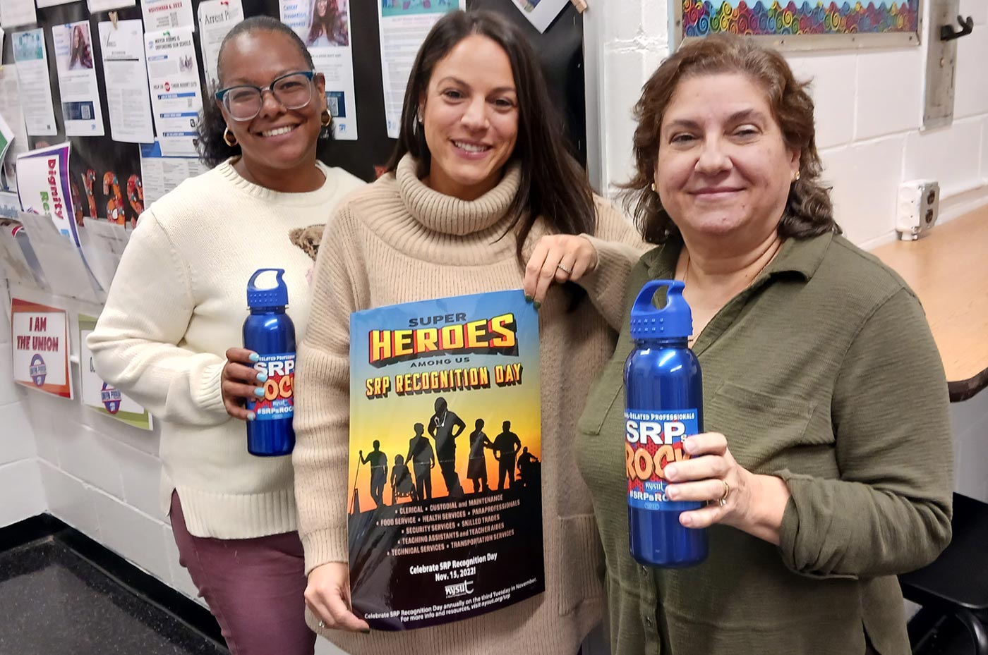 school secretaries, from left, Laura Familia, Dana Gottsegen and Wendy Guardion, stand together smiling, Laura Familia and Wendy Guardion hold water bottles with SRP Rocks stickers, while Dana Gottsegen holds a poster that reads "Super Heroes Among Us: SRP Recognition Day"