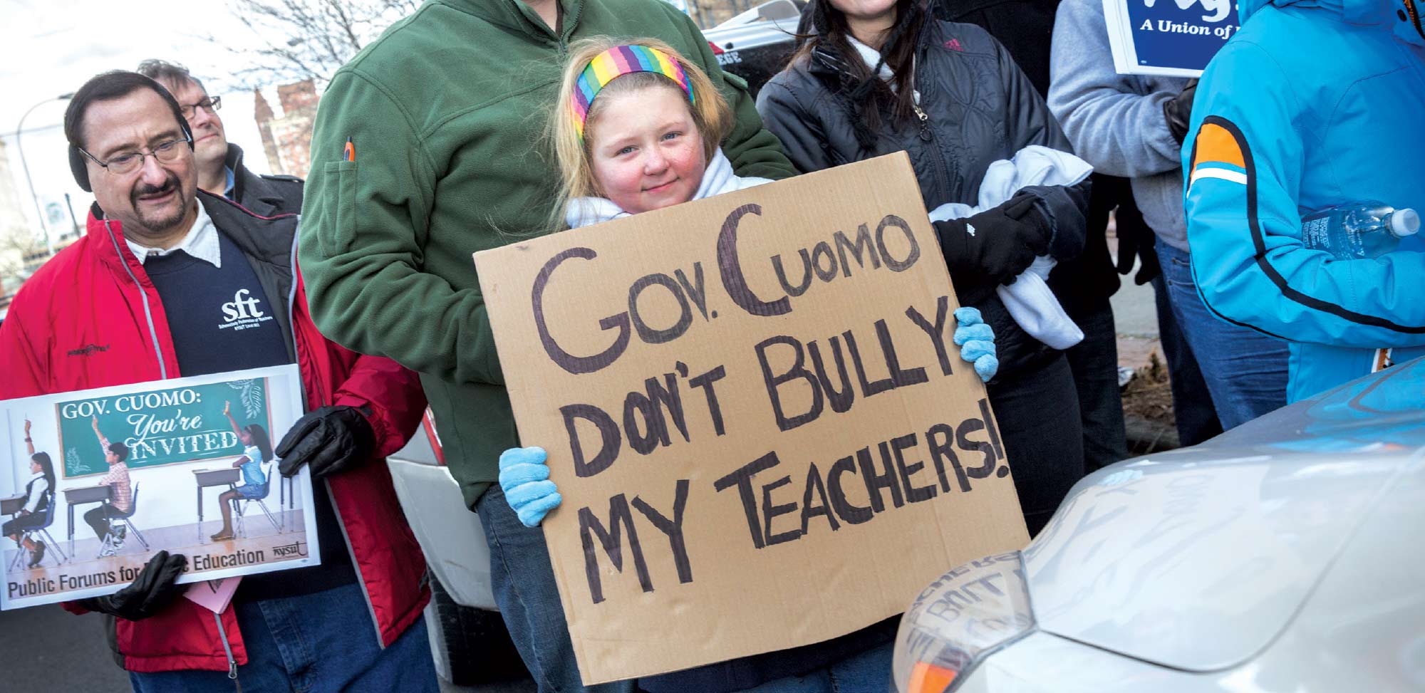 Close-up photograph perspective of a child holding a cardboard sign showing "Gov. Cuomo Don't Bully My Teachers" as she smiles in her cold weather attire while there are other people nearby her