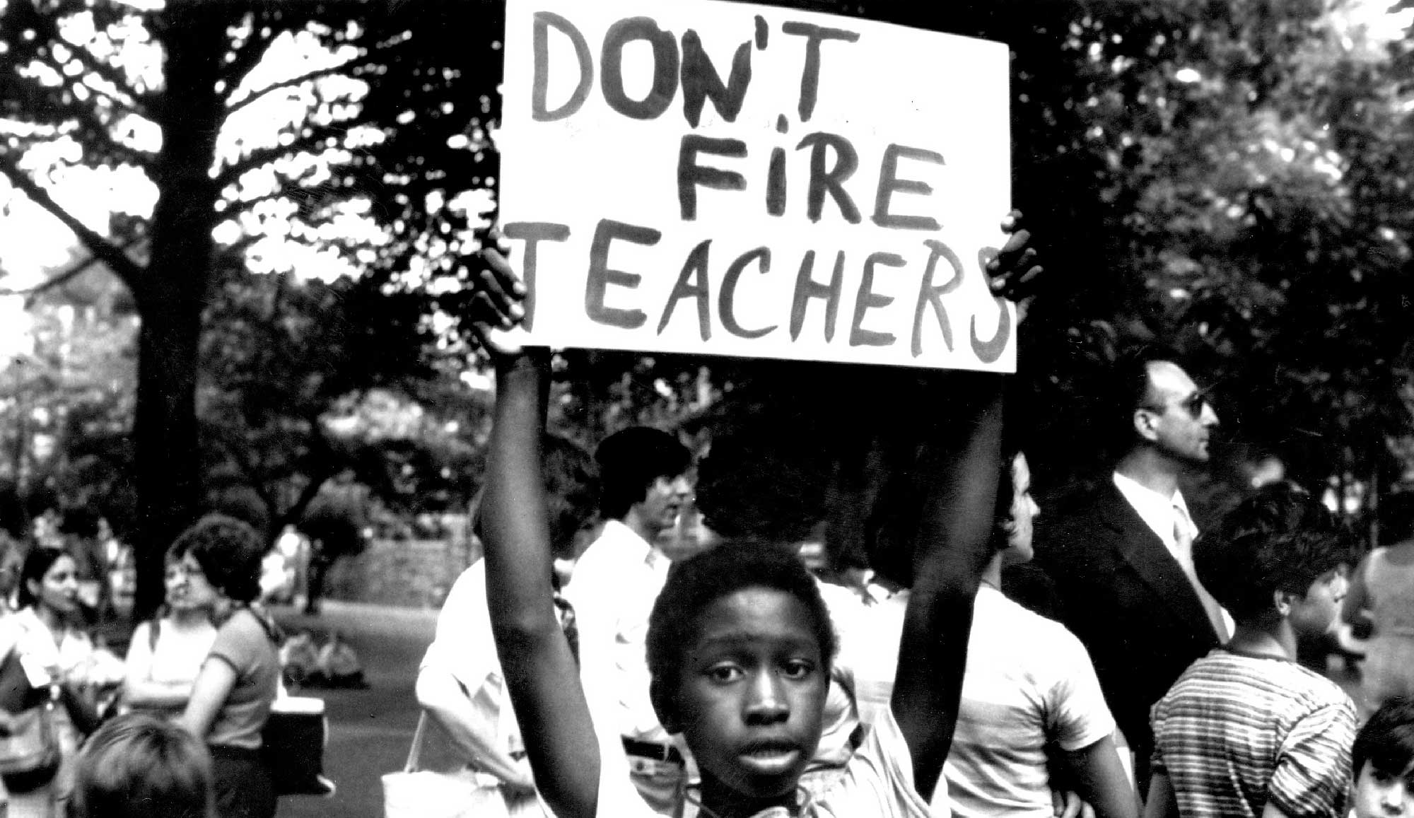 Black and white photograph close-up perspective of an African-American child holding up a sign that shows "Don't Fire Teachers" while there are other people nearby him somewhere outside
