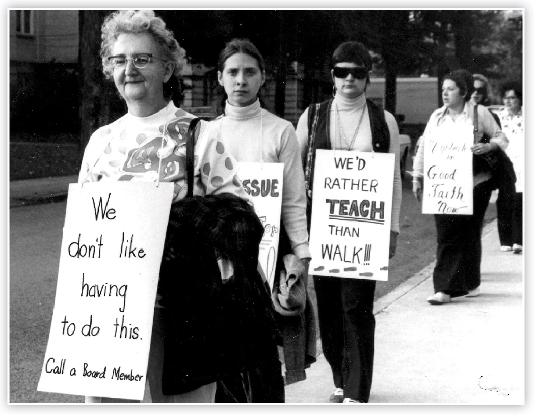 Black and white photograph close-up perspective of six women (Oneonta teachers) walking as they are holding signs with powerful statements symbolizing they are on strike in 1975
