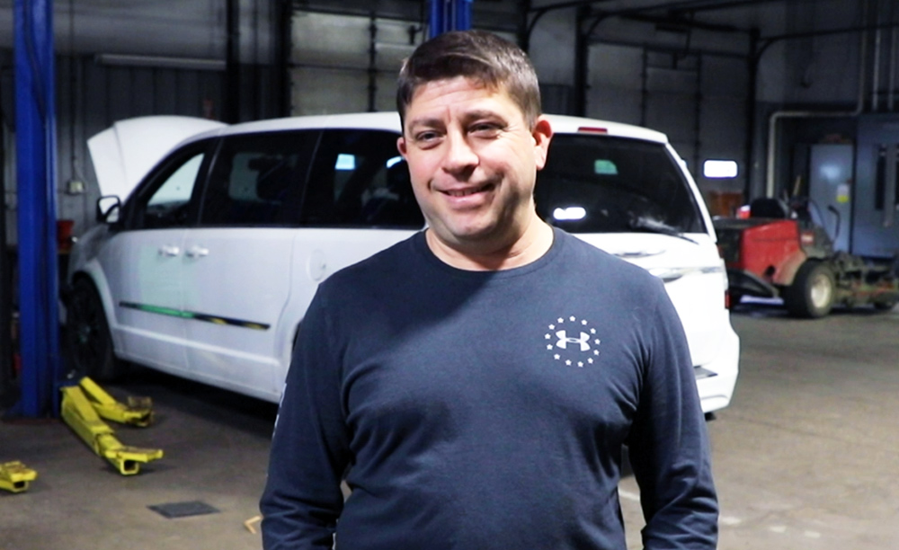 Portrait photograph of Vincent LaVerdi smiling in a navy blue Under Armour long-sleeve t-shirt as he is poses for a picture behind a white van being worked on inside an auto mechanic shop