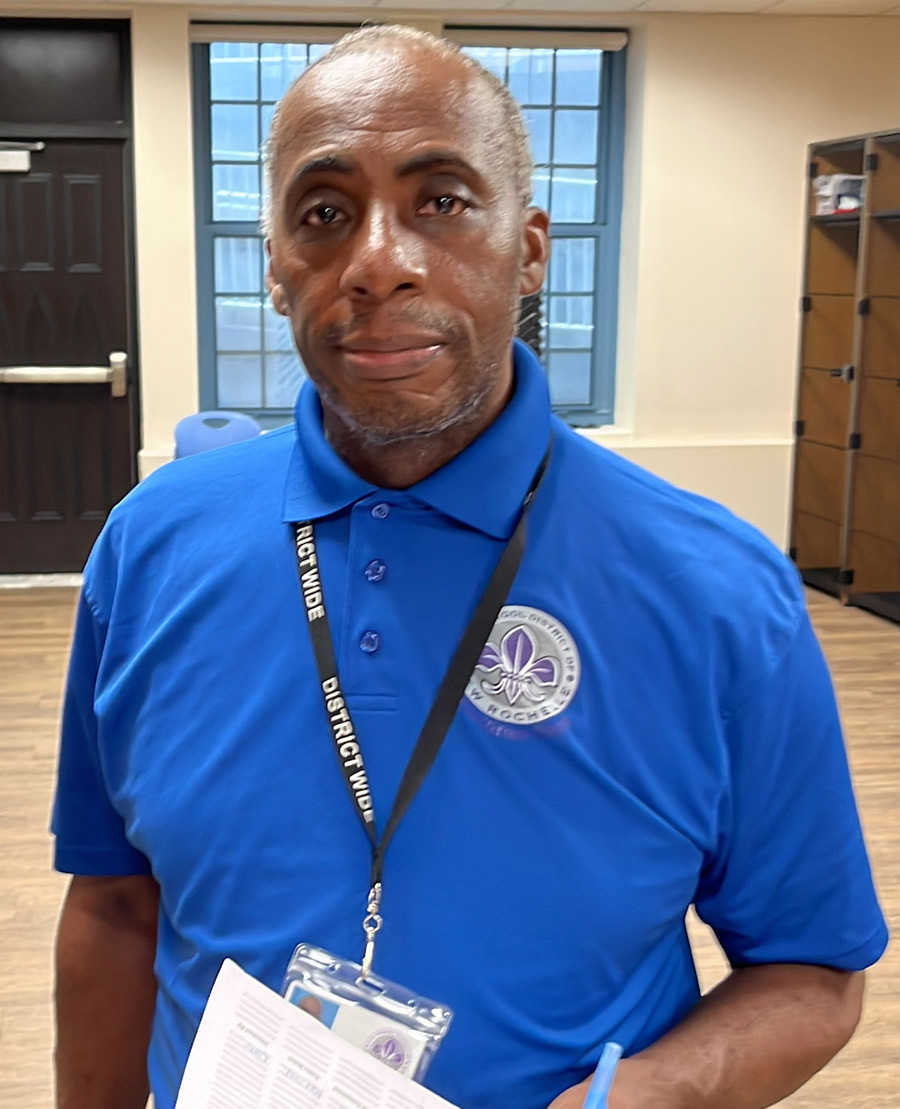 Portrait photograph of David Mills grinning in a blue buttoned-up polo top with the New Rochelle School District logo displayed on the polo top and his black lanyard/chrome badge around his neck while he holds a blue pen and white paper
