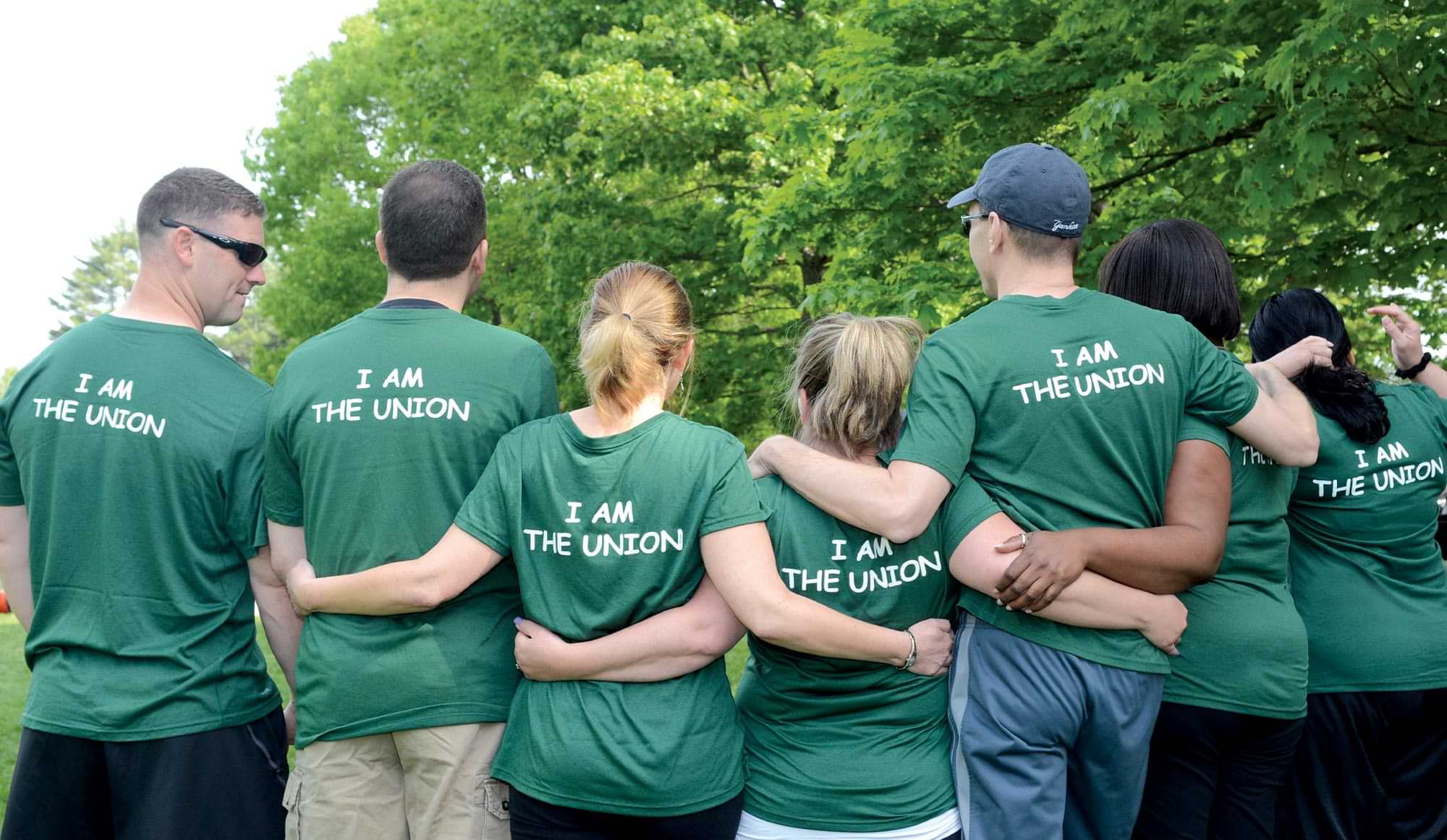 back view of a group of adults standing close, some with their arms around each other, all wearing the same green shirt that reads I am the Union in the back