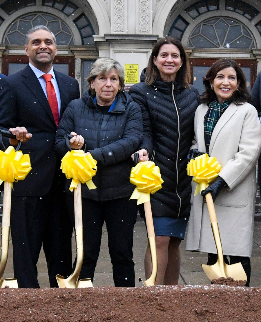 A man in a suit and three women in coats, standing with gold shovels that have a yellow bow on them. A pile of dirt in front of them and part of a building behind them.