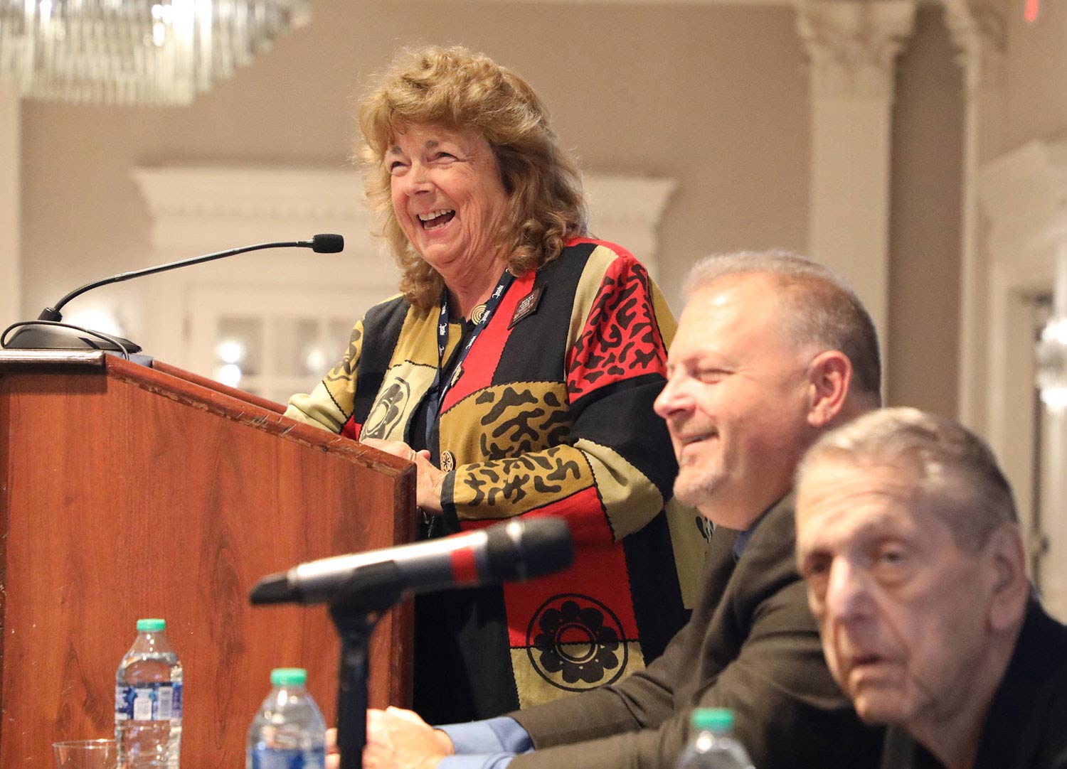 Florence McCue speaking at a podium with two men sitting in the foreground