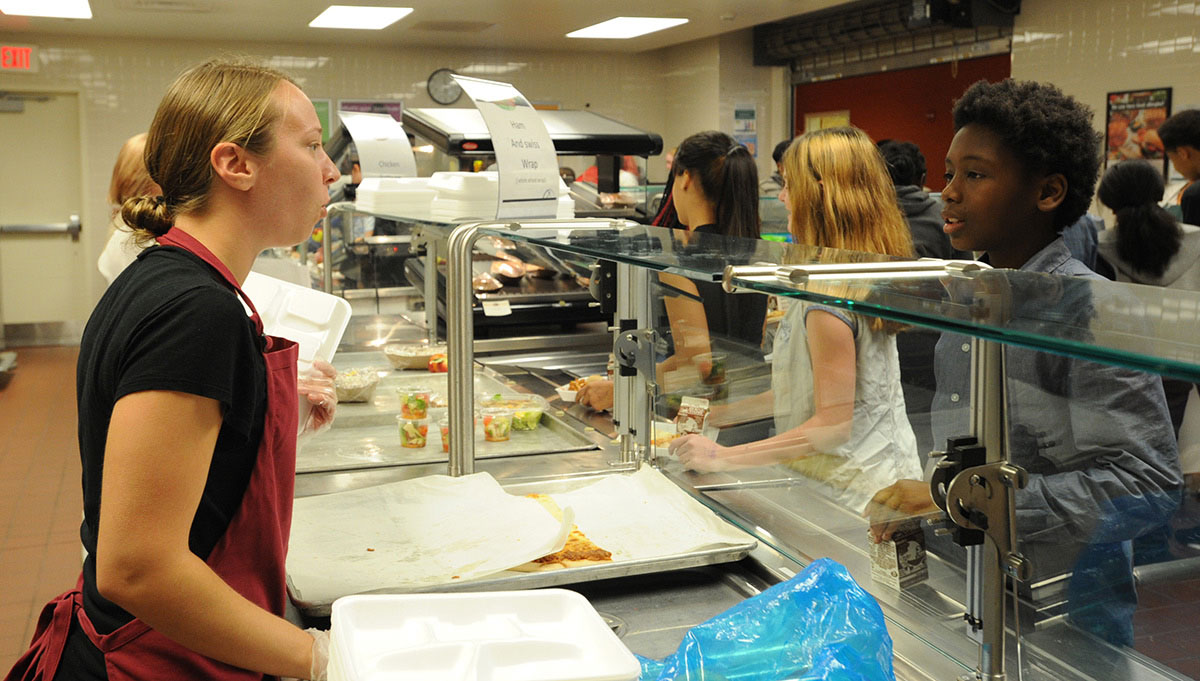 Young woman in red apron behind a lunch counter, serving lunch to students in cafeteria