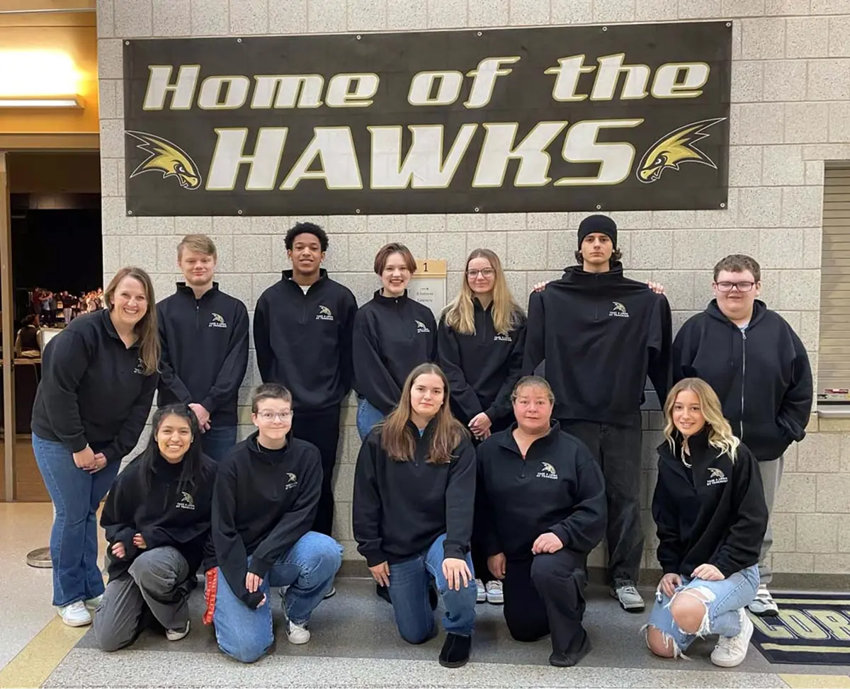 Portrait close-up photo view of Corning TA member Megan Plate smiling for a group photo with other students and another teacher in front of a wall that shows a banner displayed on the wall labled as Home of the HAWKS with the school logo on both sides of the word HAWKS