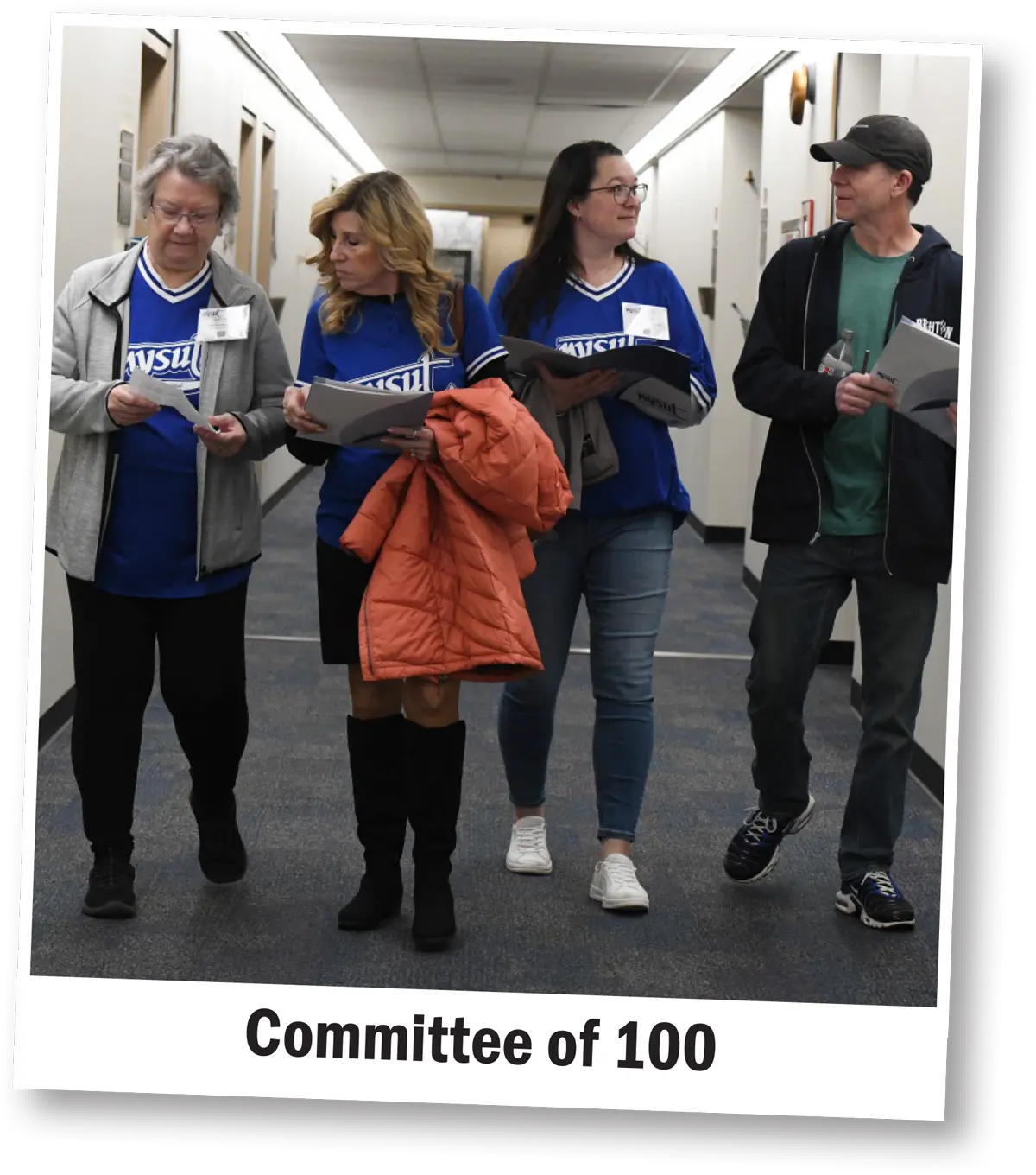 four members of the Committee of 100 walk down a hall and hold papers and folders while deep in discussion
