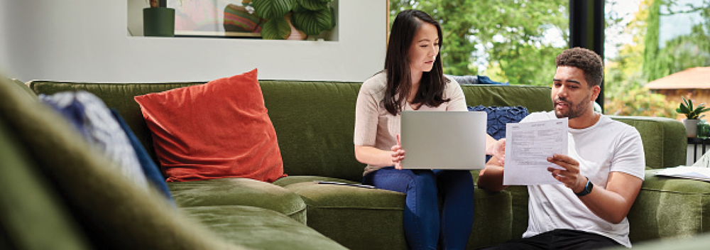 Two people on a green sofa with a laptop and papers in a living room.