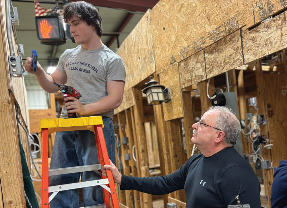 A young man on a stepladder using tools, with an older man observing in a construction setting.