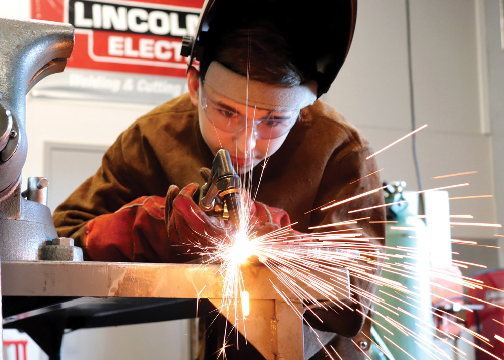 A person welding metal with sparks flying, wearing a welding helmet, protective goggles, and gloves.