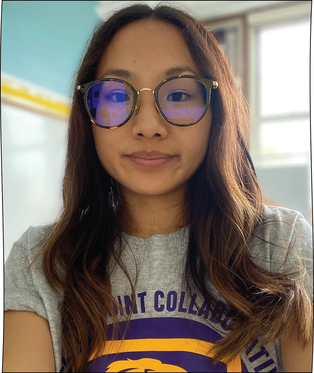 A close-up shot shows a young woman with long, wavy brown hair and light brown skin, wearing round glasses with a tortoiseshell frame. She is looking directly at the camera with a neutral expression, and she is wearing a gray t-shirt with purple and gold lettering and designs.