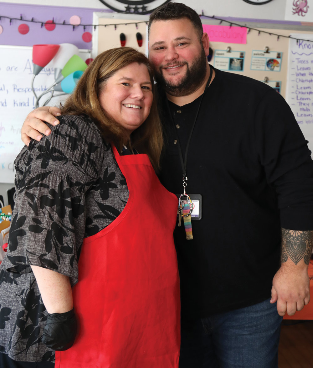 A smiling woman with shoulder-length brown hair, wearing a red apron, stands closely with a bearded man in a black long-sleeved shirt. They are in a classroom setting with colorful decorations and a whiteboard visible in the background.