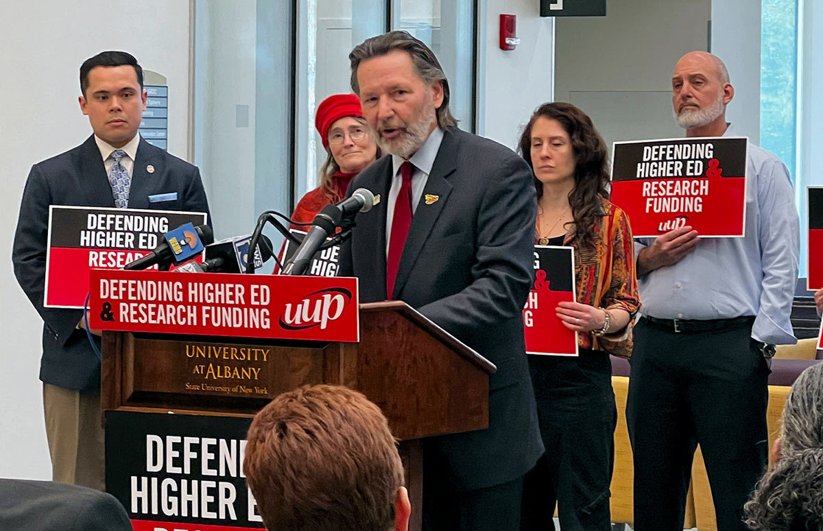 UUP President Frederick E. Kowal stands speaking into a mic'd podium at a press conference, the podium is affixed with "Defending Higher Ed & Research Funding" signs, with similar signs being held by the few people standing behind Frederick E. Kowal