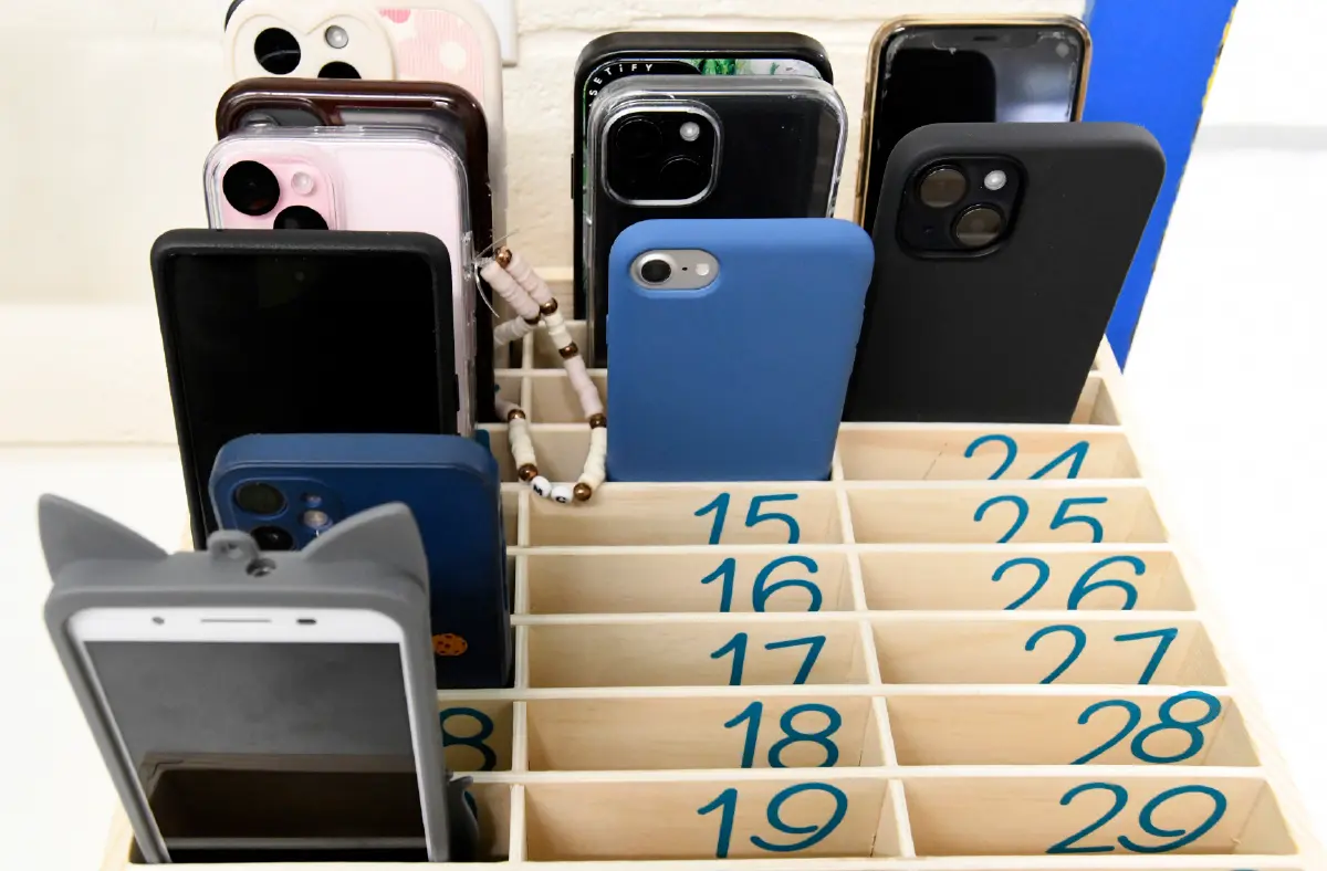 A collection of smartphones, many in cases, neatly organized in a wooden tiered holder with numbered slots.