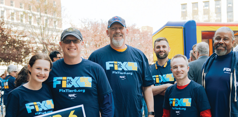 A group of six people wearing blue "Fix Tier 6" T-shirts, posing outdoors with buildings and an inflatable structure in the background.