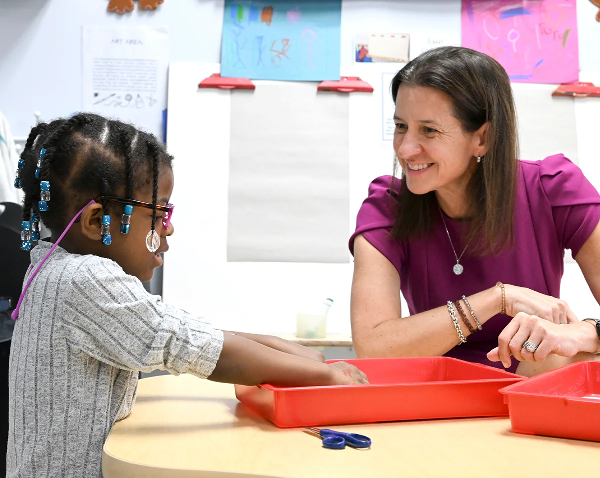 NYSUT President Melinda Person smiling at a young student with glasses and beaded braids, as they engage in a classroom activity with red trays on a wooden table.