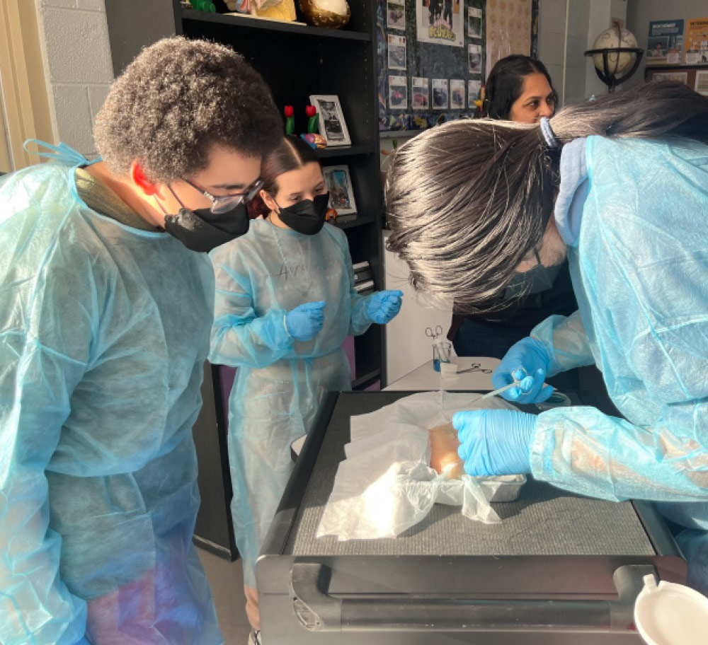 Three students wearing light blue surgical gowns and blue gloves are gathered around a table. One person on the right is actively working with tools on what appears to be a practice surgical pad during a lab