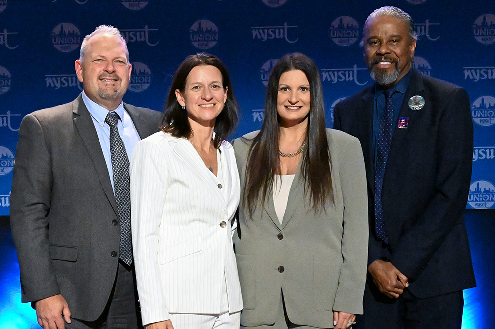 A group of four adults smiling at the camera in front of a blue backdrop with the NYSUT Union logo.