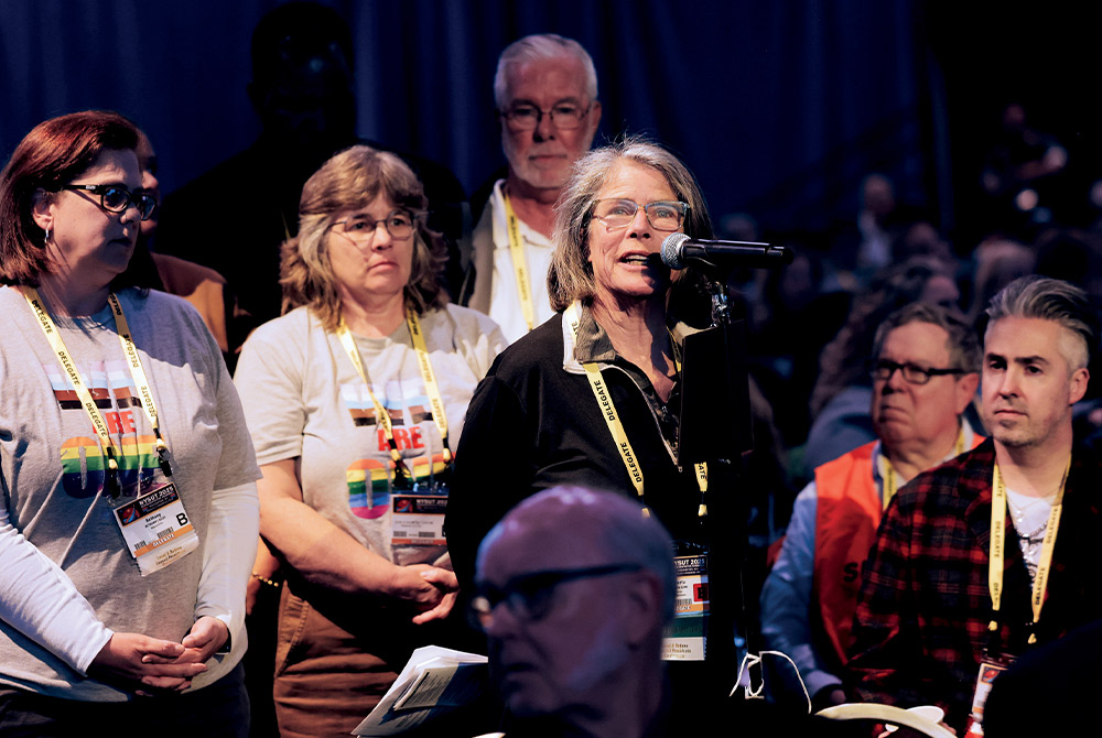 A woman with shoulder-length gray hair and glasses speaks into a microphone, surrounded by other people in an audience.