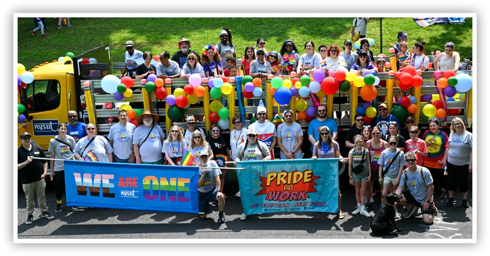 A large group of people stand around a decorated truck with balloons and banners, some holding a "WE ARE ONE" banner and a "PRIDE @ WORK" banner.