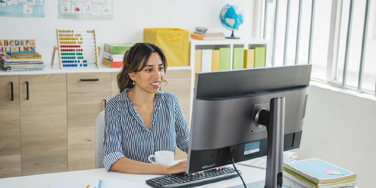 A professional woman with dark hair in a striped shirt sits at a desk, looking at a computer monitor and typing, with a mug nearby. The setting appears to be an office or classroom with cabinets, books, and a globe visible.