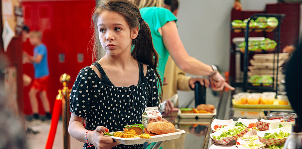 A young girl in a school cafeteria line holds a lunch tray with a sandwich, fries, and a milk carton.