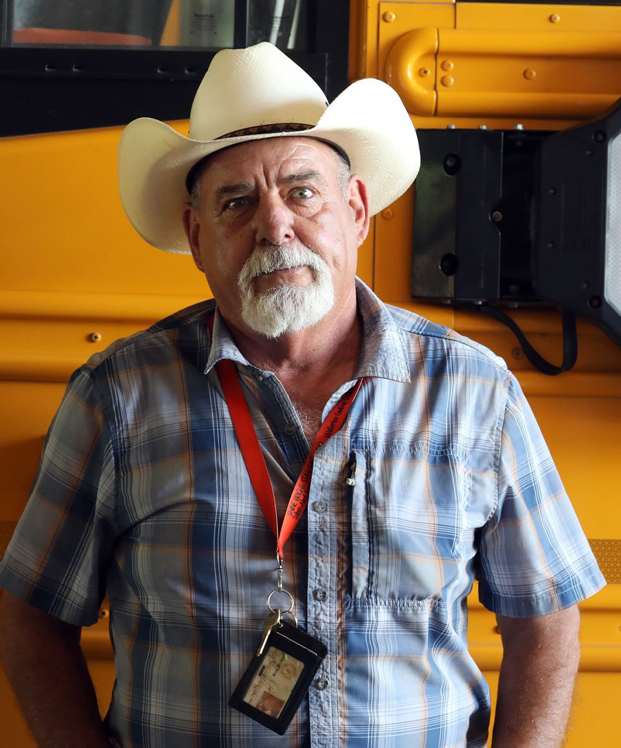 Headshot of Rick Leisenring, an older man in a cowboy hat and plaid shirt, possibly a school bus driver, standing in front of a yellow bus.