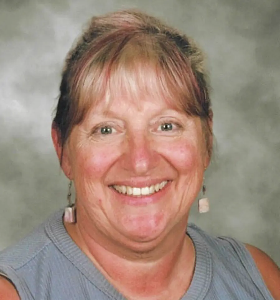 A studio portrait of Katy Smith, a smiling woman with blonde hair and a fringe; She is wearing a light blue sleeveless top and small drop earrings, set against a mottled gray and white background