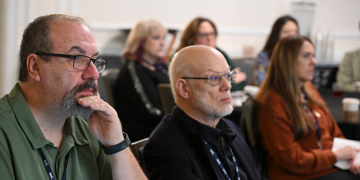 A close-up, side-angle view of several people sitting at a table during a meeting. A man in the foreground with a beard and glasses rests his chin on his hand, looking forward with a serious, contemplative expression.