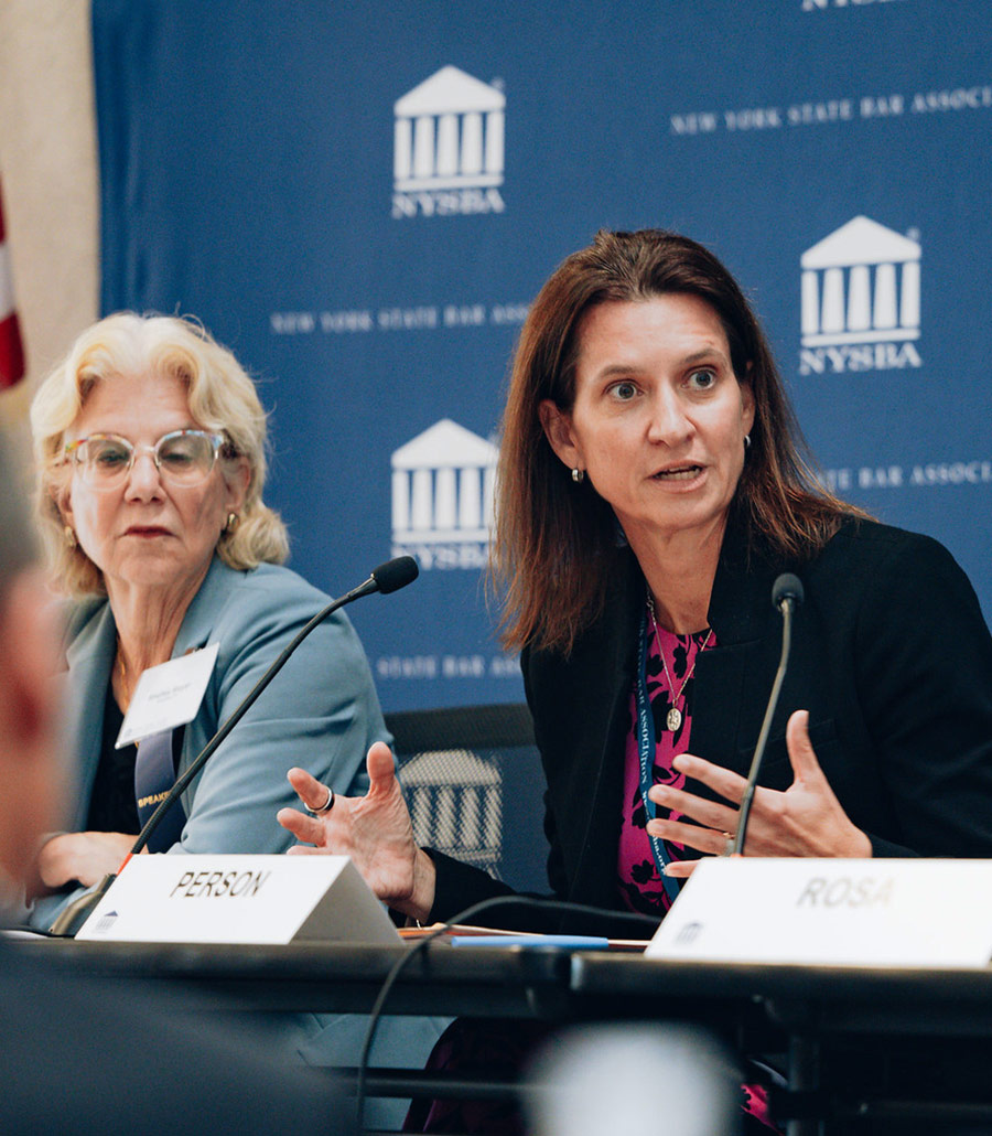Melinda Person gestures expressively while speaking into a microphone. Shelley Mayer sits to her left, listening attentively. They are positioned in front of a blue backdrop with repeated white NYSBA logos.