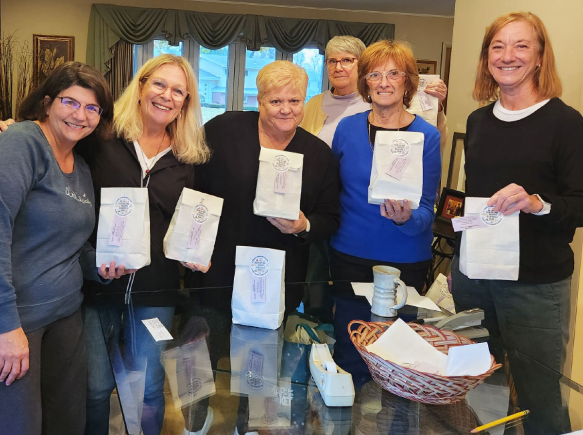 Women smiling and holding up white gift bags with circular labels while standing around a glass table.