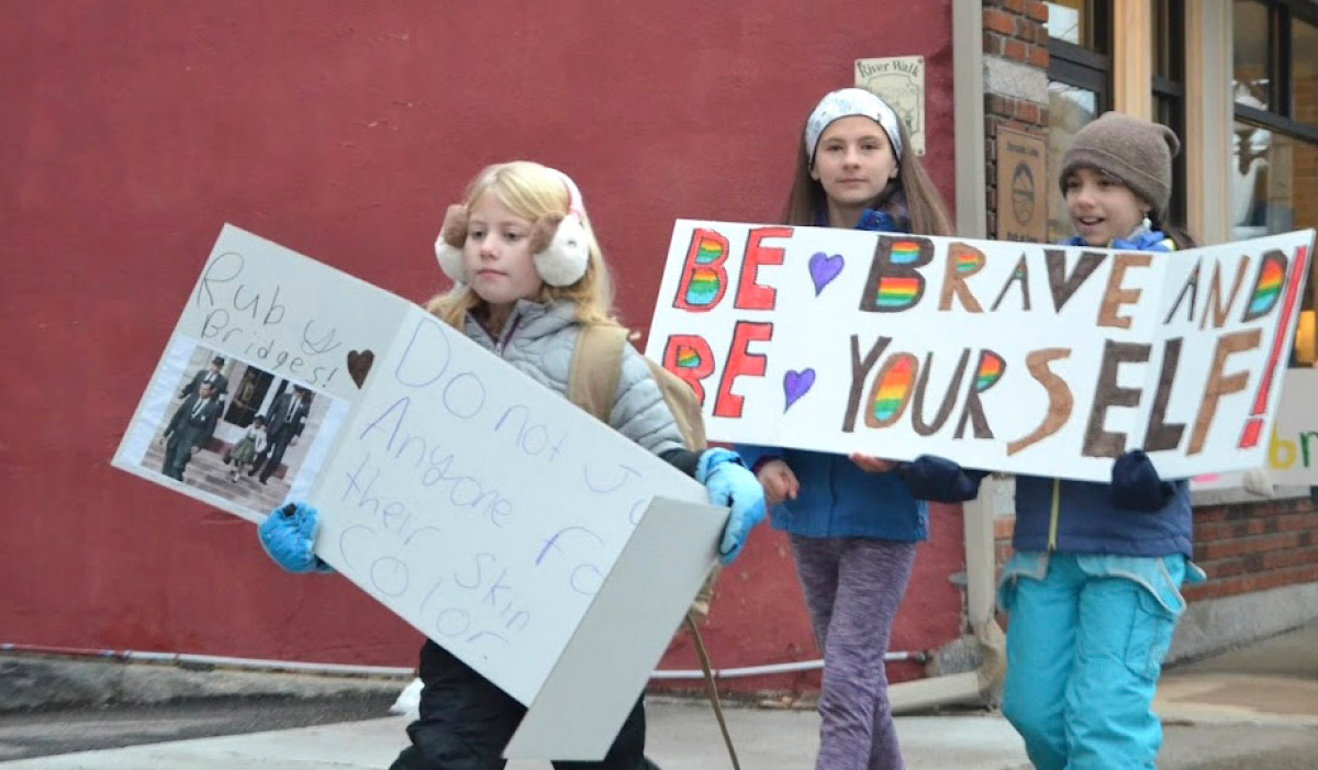 Three young girls walking outdoors holding handmade signs with messages about bravery and racial equality.