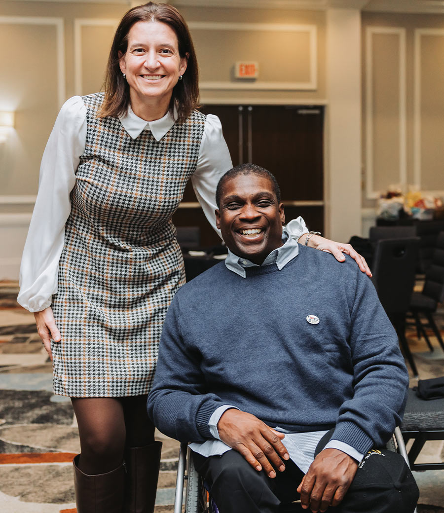 Melinda Person stands with her hand on the shoulder of a smiling James Chaney, who is seated in a wheelchair. They are both dressed in professional attire at a conference event with a blurred ballroom background.