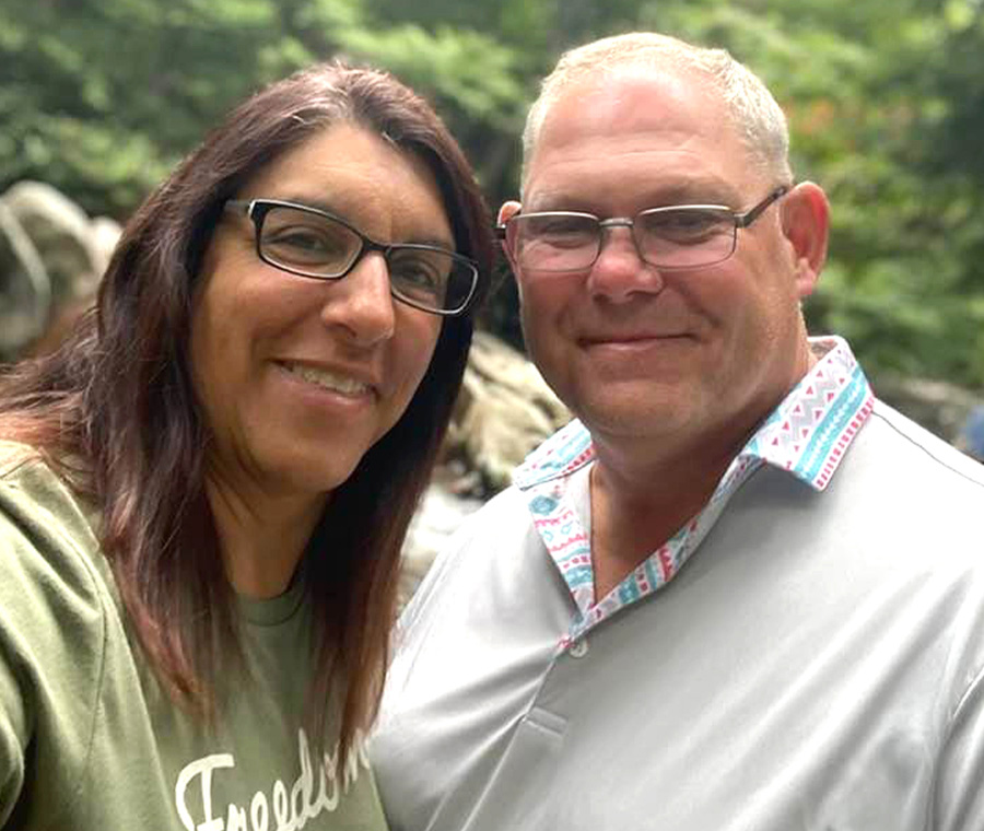 A man and a woman smile for a close-up outdoor photo. The woman has long dark hair and glasses; the man wears glasses and a light-colored polo shirt. They stand close together against a soft-focus background of lush green foliage.