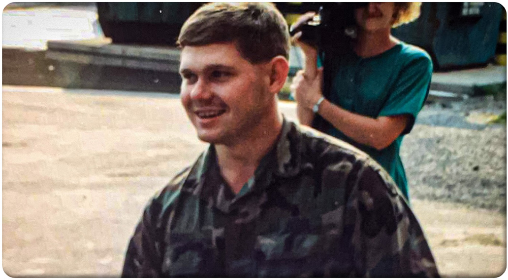 A smiling young man in a camouflage U.S. Army uniform stands outdoors and looks off to the side. The photo is a candid shot with a slightly blurred background.