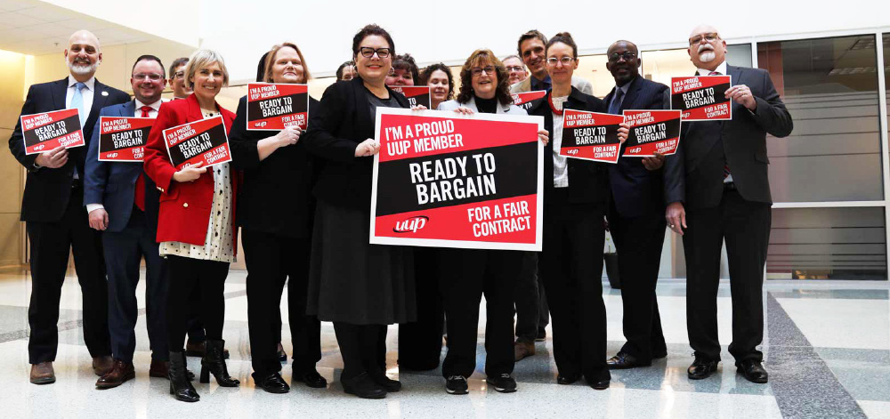 A large group of people stand together indoors holding red and white signs that read, “I'M A PROUD UUP MEMBER READY TO BARGAIN FOR A FAIR CONTRACT.”