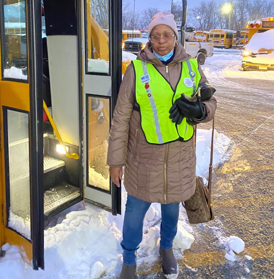 Deirdre Mostiller pictured wearing a neon green safety vest over a brown winter coat standing in the snow next to an open school bus door.