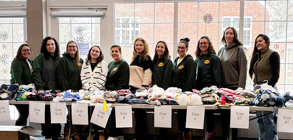 Eleven Westbury TA members stand behind a long table stacked with folded pajamas. Paper signs taped to the table indicate various clothing sizes. The group is positioned indoors in front of large windows overlooking a brick building.