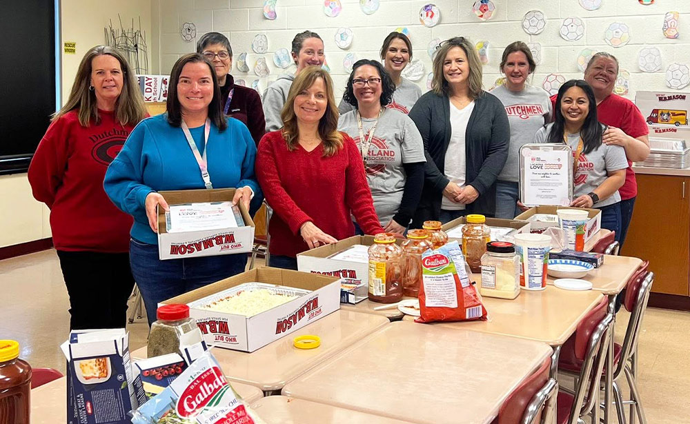 Eleven members of the Guilderland TA Social Justice Committee stand in a classroom behind desks holding pans of lasagna, jars of sauce, and bags of cheese. Student drawings of soccer balls are visible on the wall.