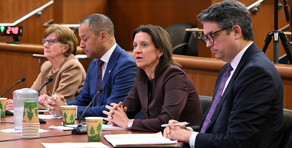 NYSUT President Melinda Person speaks at a microphone while seated at a long table with three colleagues. They are testifying before the joint Assembly and Senate Education Committees in a formal wood-paneled hearing room.