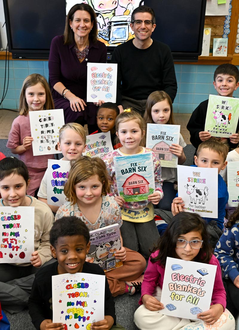 NYSUT President Melinda Person and a man sit with elementary students holding signs with slogans like “Electric Blankets For All” and “We Strike.” One child holds the book “Click, Clack, Moo: Cows That Type.”