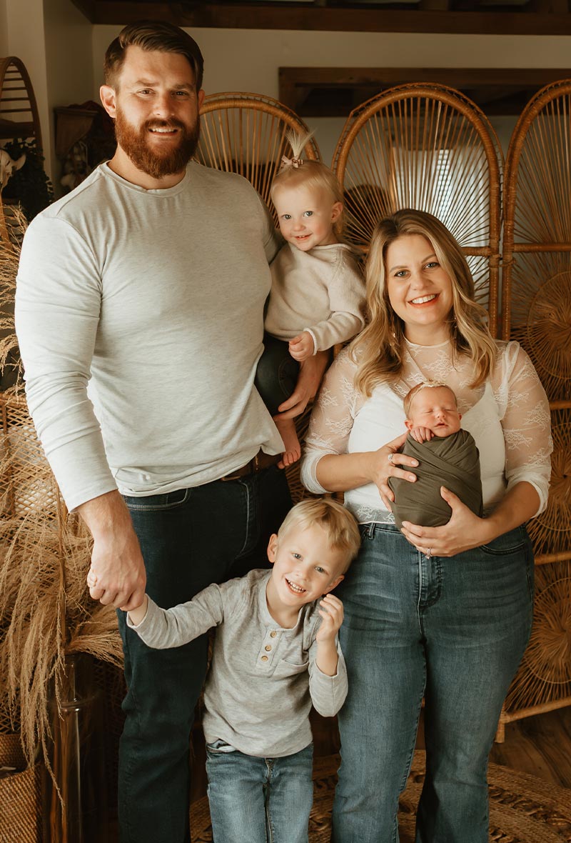 Kathryn Luczak and her husband pose for a family portrait with their three young children, including a swaddled newborn baby, in front of a decorative wicker screen.