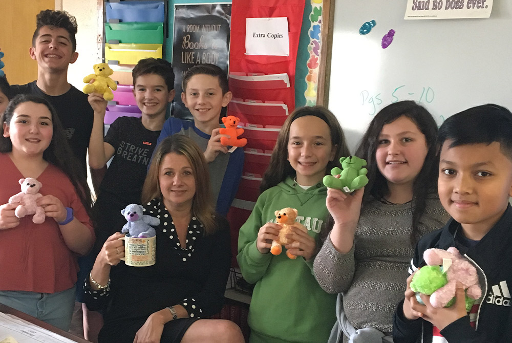 Michele Witte and eight students smile in a classroom, holding colorful "Tyler’s Teddies" in memory of her son. Witte sits centered with a blue bear in a mug, surrounded by students displaying pink, green, and orange stuffed bears.