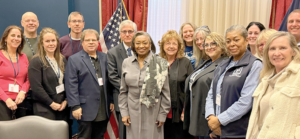 A group of people pose in a formal room with flags. Senate Majority Leader Andrea Stewart-Cousins stands in the center in a grey dress and floral scarf. A small inset image shows an outdoor event under a blue Federation of Teachers tent.