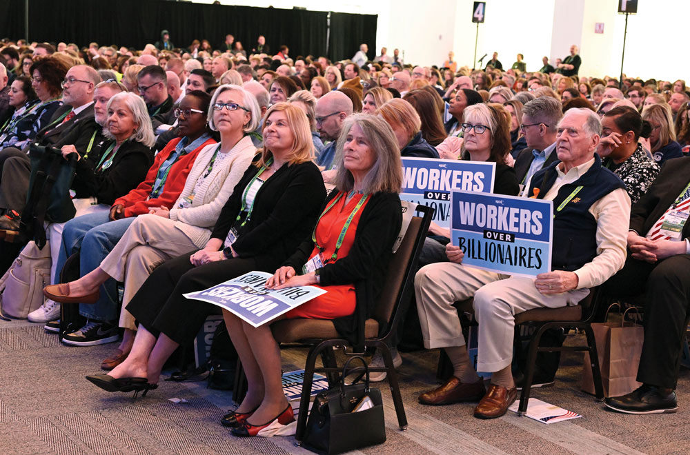 An audience at a conference listening to a speaker, with several attendees holding "Workers Over Billionaires" signs.