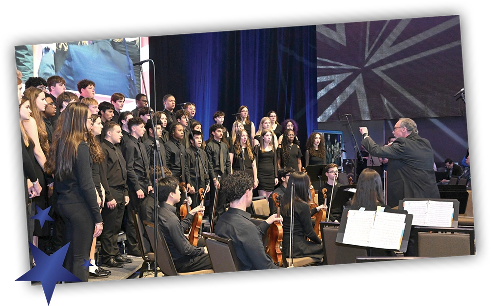 A student choir in black attire performs alongside a small orchestra, guided by a conductor on a convention stage.